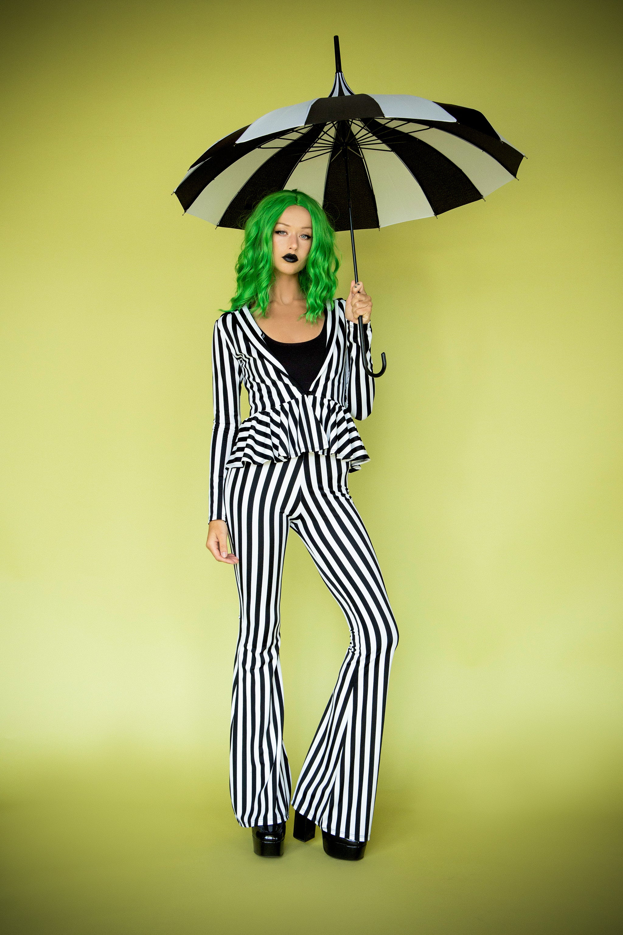 A person wearing a black and white striped suit with a jacket and bell bottoms, holding an umbrella, standing against a yellow background.